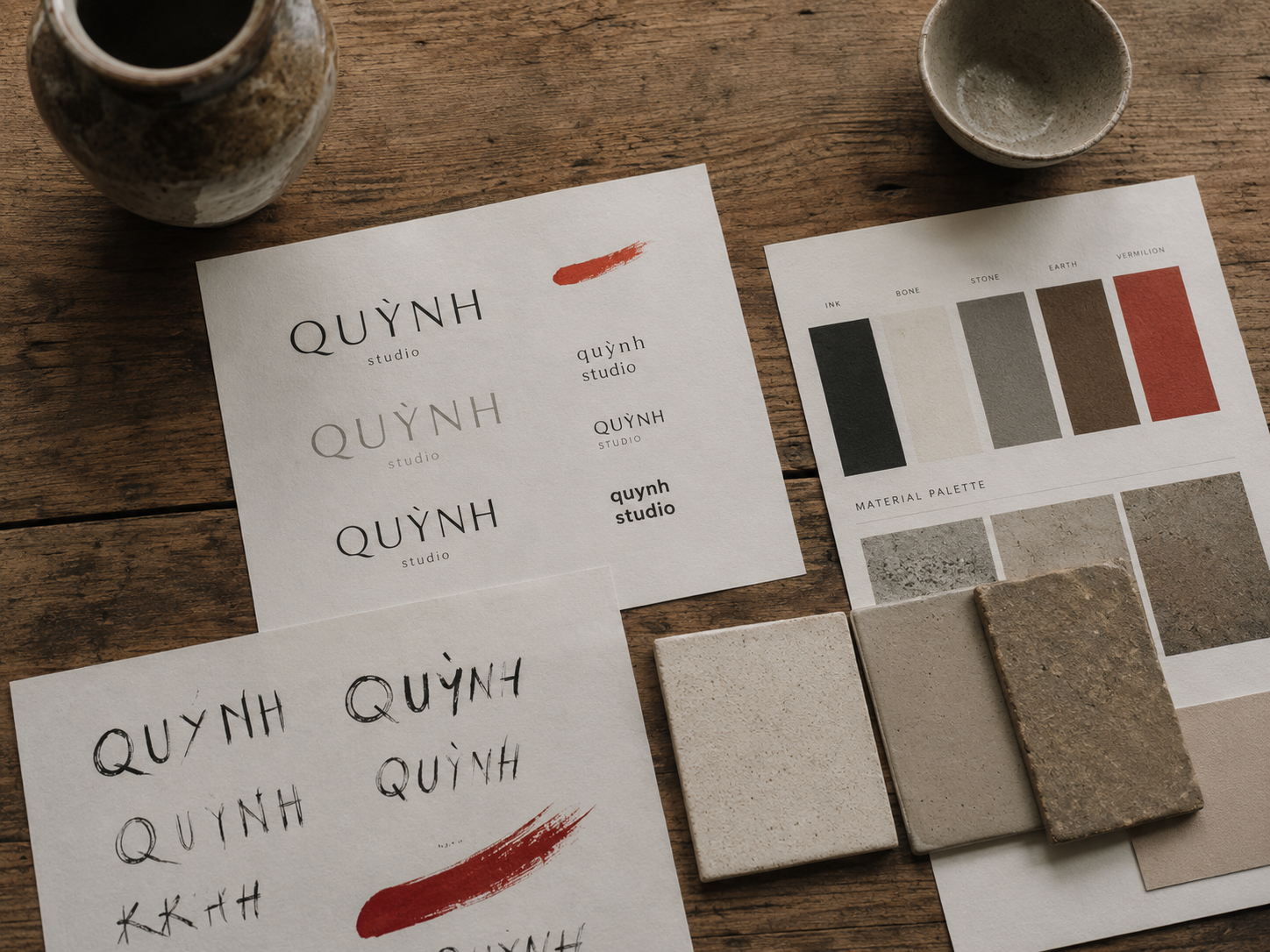 Top-down view of a printed brand-identity exploration spread on a raw timber drafting table, typographic experiments, colour swatches, ceramic-tile samples, single vermillion brush mark